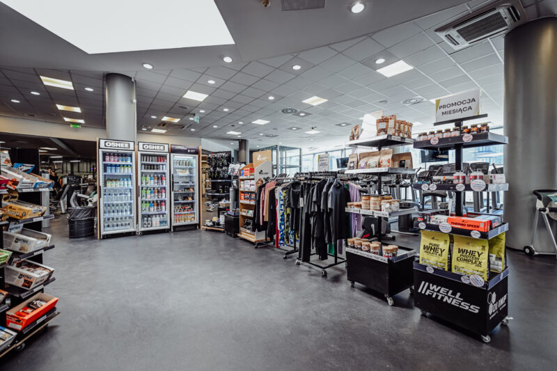 Strefa przy recepcji Well Fitness Legnica Galeria Ferio – produkty proteinowe, napoje i odzież sportowa.