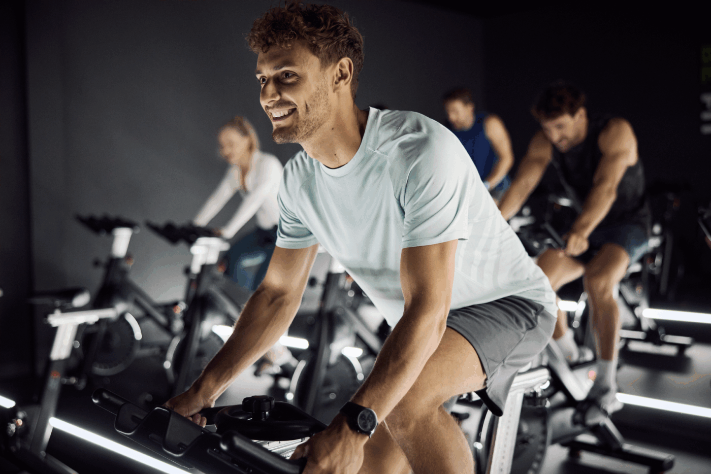 Uczestnicy zajęć Indoor Cycling w klubie fitness ćwiczący na rowerach stacjonarnych, uśmiechnięty mężczyzna na pierwszym planie.