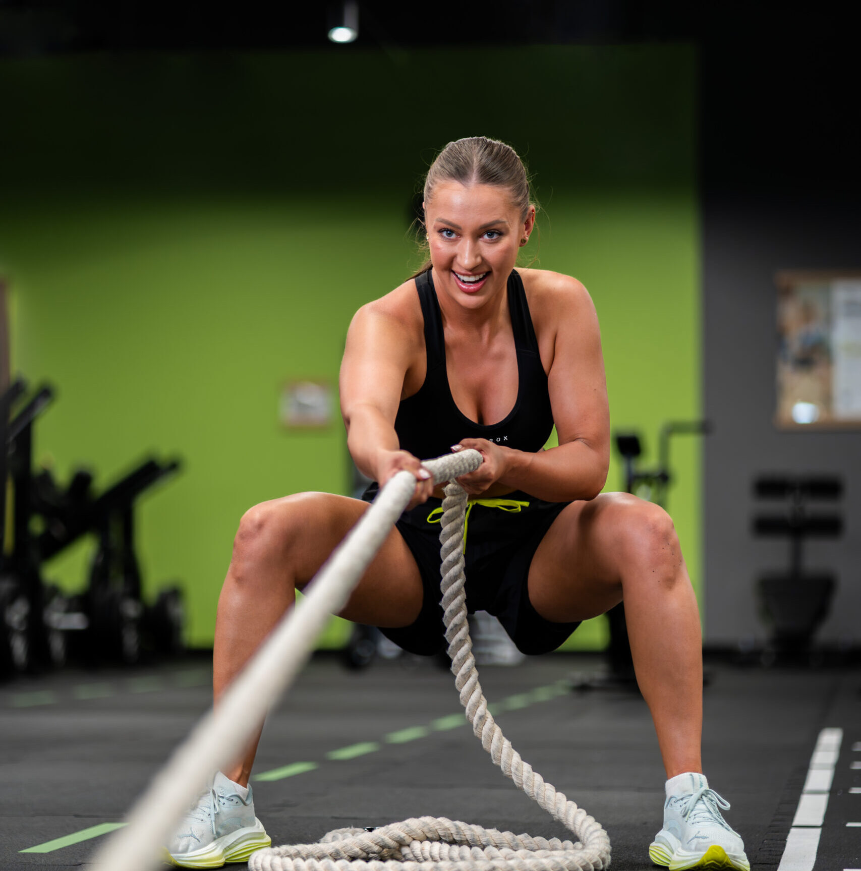Uśmiechnięta kobieta podczas wymagającego treningu HYROX w Well Fitness, dynamicznie przeciągająca linę, w nowoczesnym wnętrzu klubu.