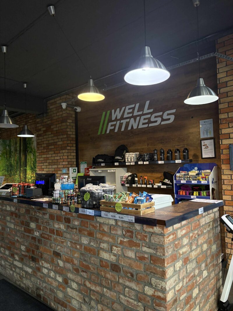 Recepcja klubu Well Fitness z obsługą i stanowiskiem rejestracji.