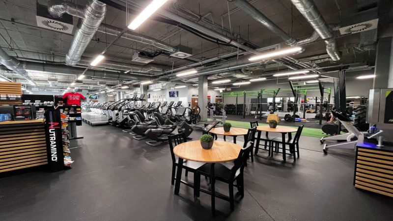 Strefa do odpoczynku. Na środku otwartej sali stoją stoły oraz krzesła, w tle widać drążki i maszyny do cardio. Po lewej stronie znajduje się recepcja.
