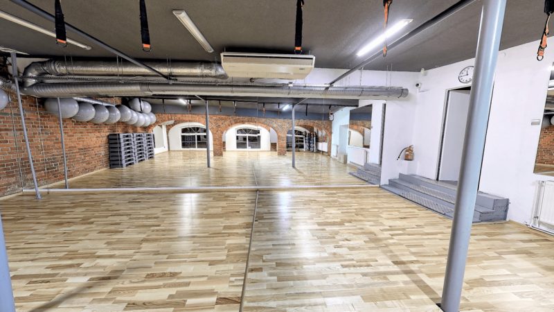Sala fitness do zajęć grupowych z lustrem. Pod ścianą po prawej stronie stoją stepery.