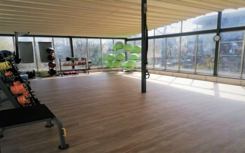 Przeszklona sala fitness do zajęć grupowych. Pod oknem oraz ścianą stoją akcesroia do ćwiczeń.