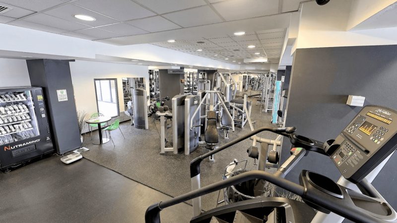 Sala z maszynami do ćwiczeń określonych grup mięśni. Po lewej stronie strefa chillu - stoliki i krzesła.