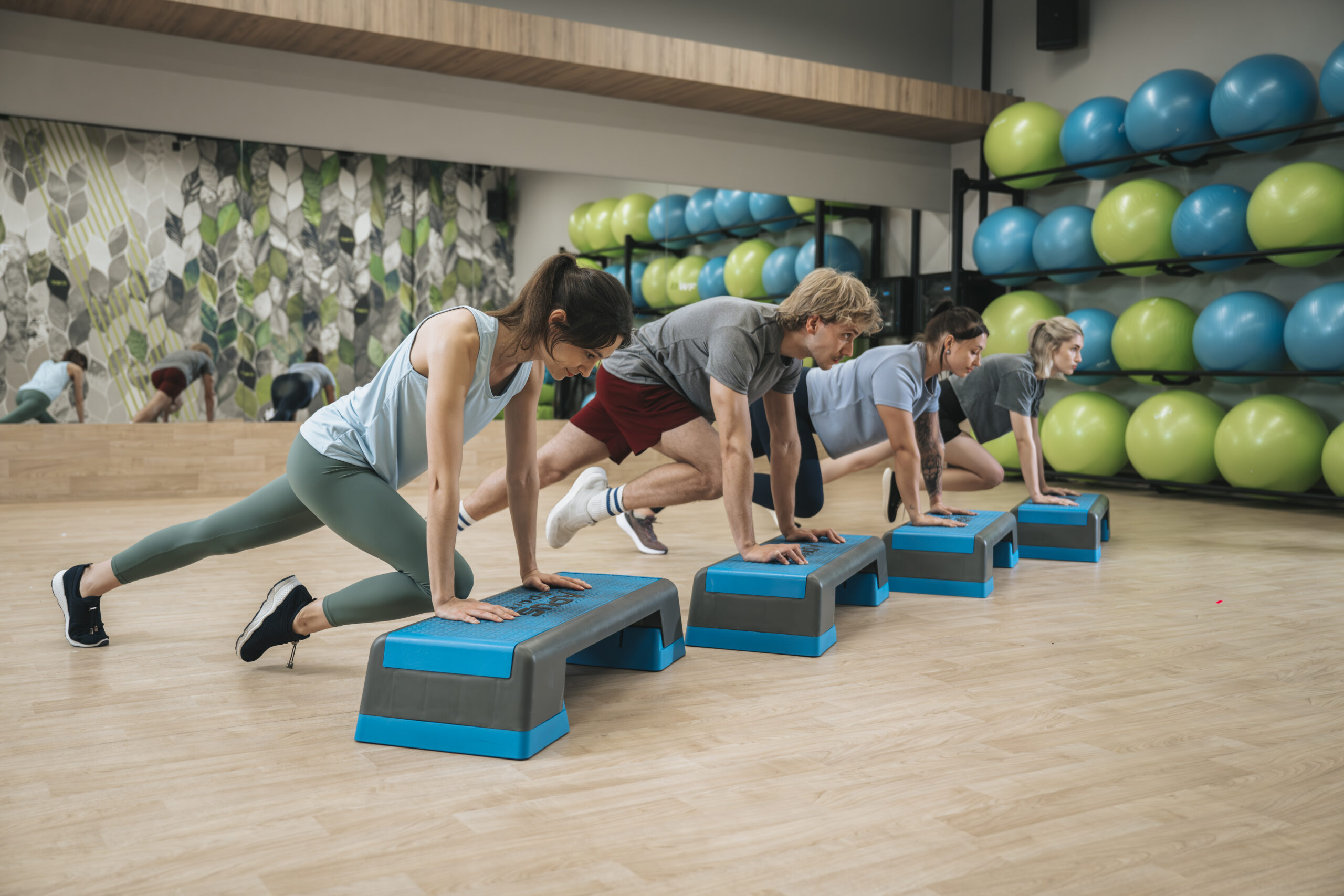 Grupa osób ćwicząca z użyciem stepów w sali fitness z piłkami gimnastycznymi w tle