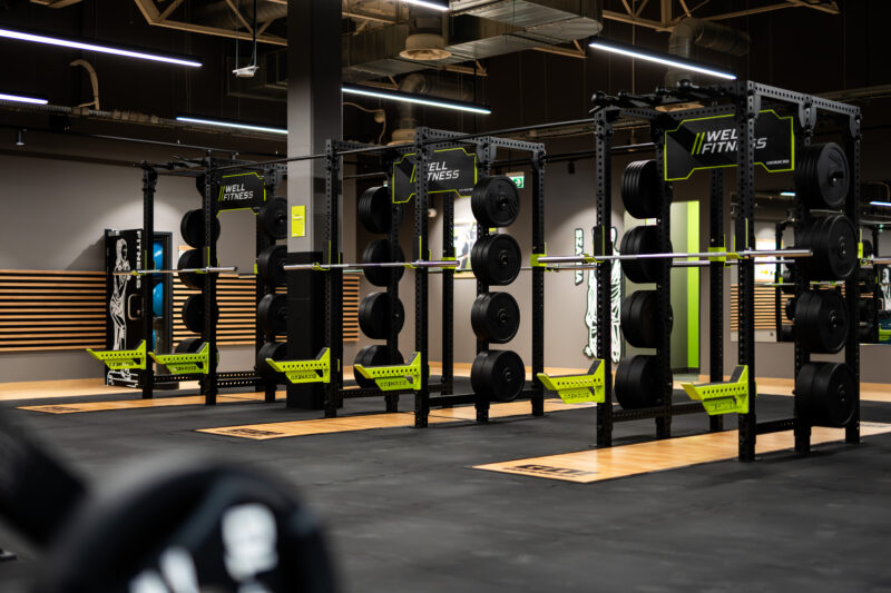 Strefa wolnych ciężarów w siłowni Well Fitness z maszynami do przysiadów i talerzami obciążeniowymi