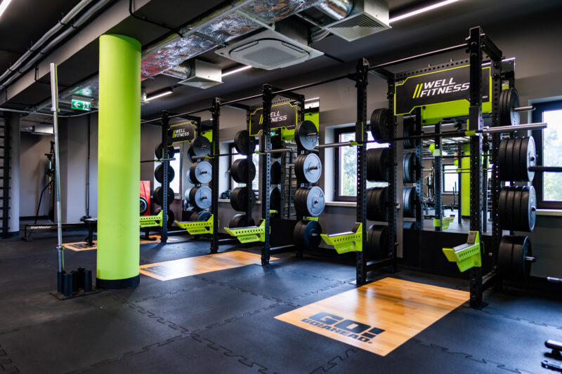 Stanowiska do treningu siłowego w siłowni Well Fitness, z gumową podłogą i platformami drewnianymi.
