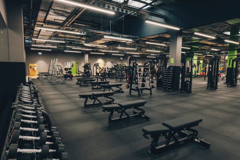 Strefa wolnych ciężarów – alt="Strefa wolnych ciężarów z ławkami prostymi, hantlami i maszynami do treningu siłowego w Well Fitness Gdańsk Oliwa