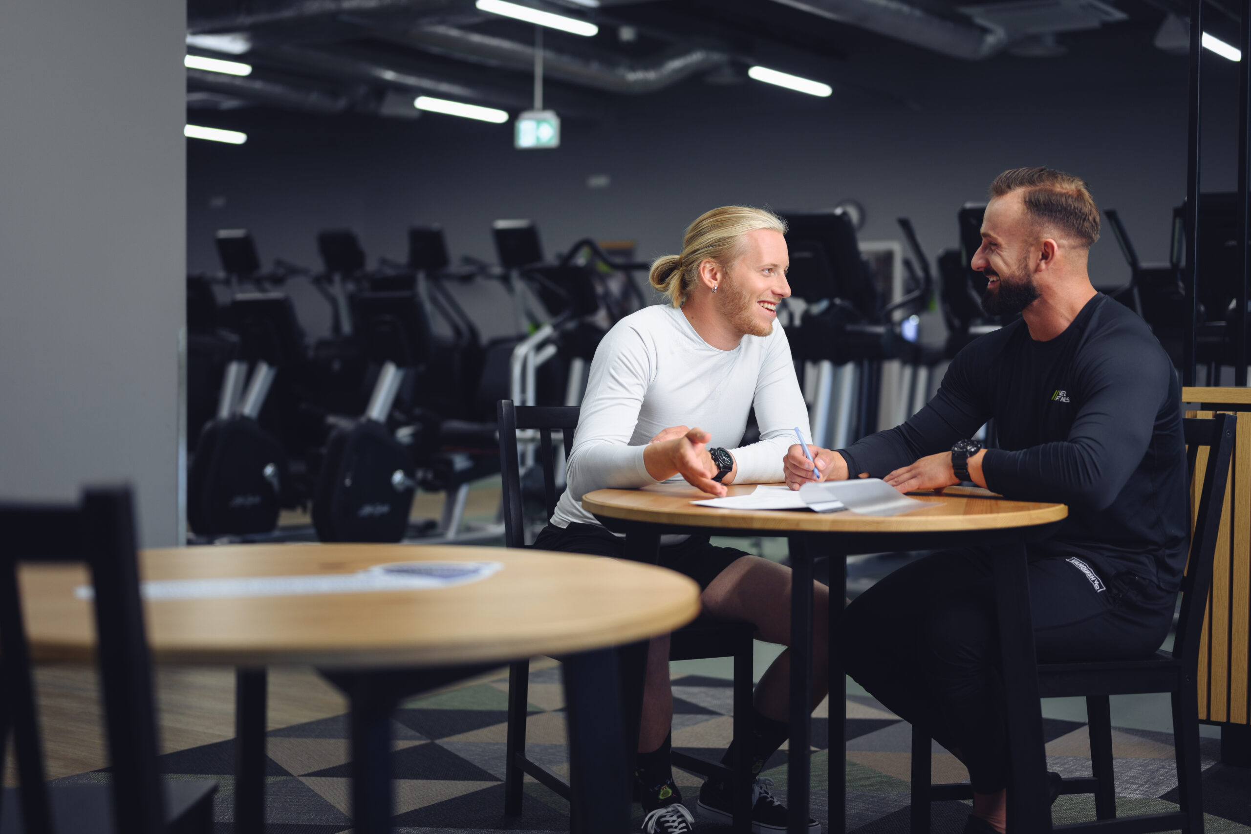 Dwóch mężczyzn podczas konsultacji z trenerem personalnym w siłowni Well Fitness - rozmowa przy stoliku, w tle sprzęt cardio.
