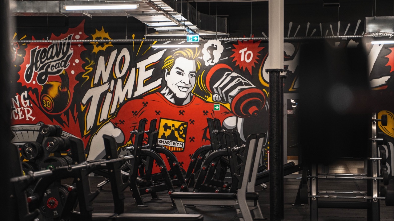 Wnętrze siłowni z widocznymi ławkami do ćwiczeń. W tle duży, kolorowy mural przedstawiający umięśnionego mężczyznę z hantlem i napisem „NO TIME”. Na koszulce postaci widnieje logo Smart Gym.