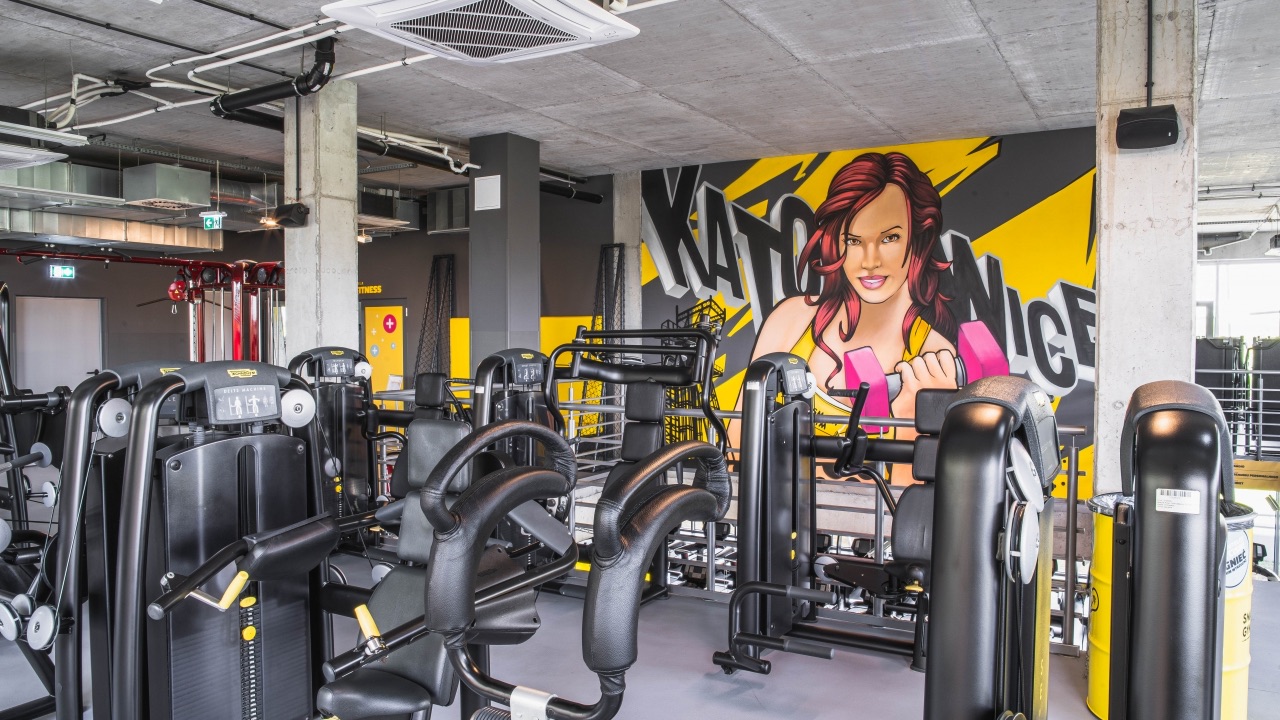 Nowoczesna sala treningowa z maszynami cardio i siłowymi marki Technogym. Na ścianie w tle widoczny komiksowy mural z wizerunkiem kobiety trzymającej hantel oraz napisem „KATOWICE”