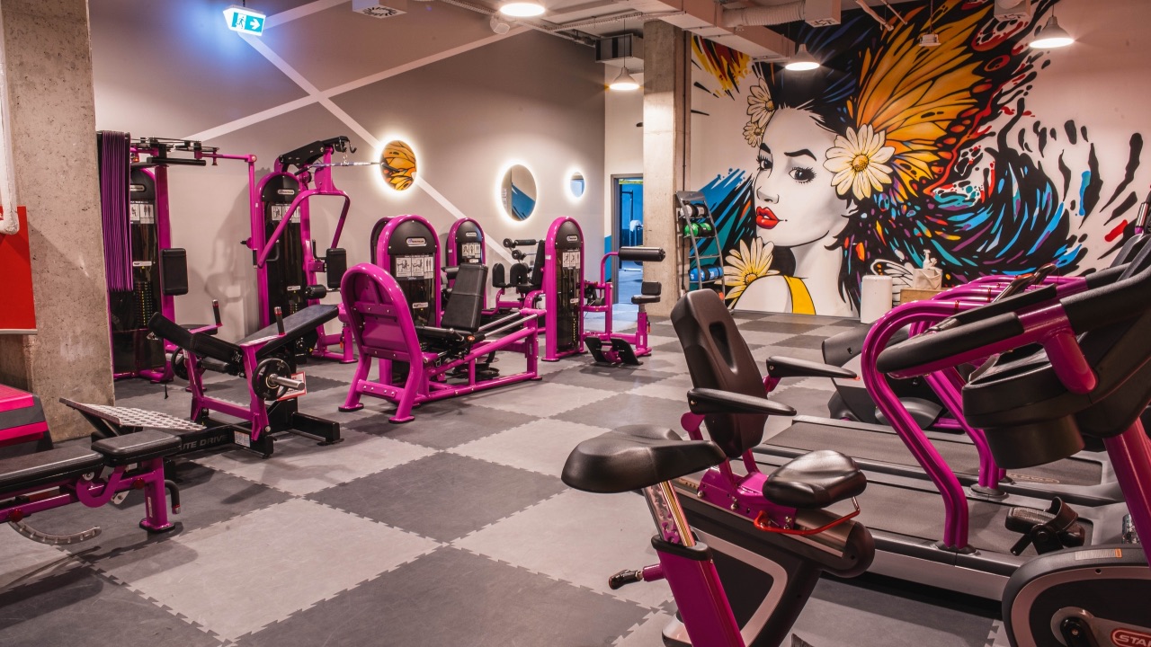 Zdjęcie przedstawia wnętrze nowoczesnej, przestronnej siłowni Smart Gym Zabrze Arena. W pomieszczeniu o industrialnym charakterze rozmieszczono liczne maszyny do treningu siłowego oraz strefę cardio z rowerami stacjonarnymi i bieżniami w intensywnym, fioletowym kolorze.