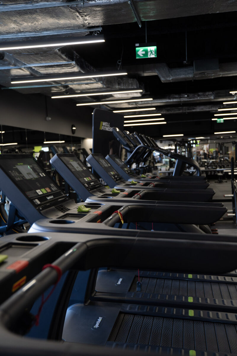 Perspektywa rzędu maszyn cardio marki Matrix, ukazująca rytmiczne ułożenie czarnych uchwytów na tle industrialnego, ciemnego sufitu z liniowym oświetleniem.