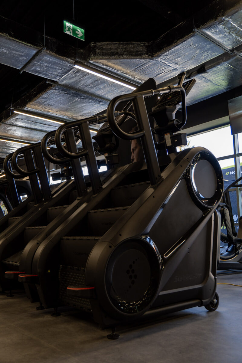 Detale konstrukcyjne maszyn do cardio typu schody, widoczne na tle jasnego okna i industrialnego sufitu klubu fitness.