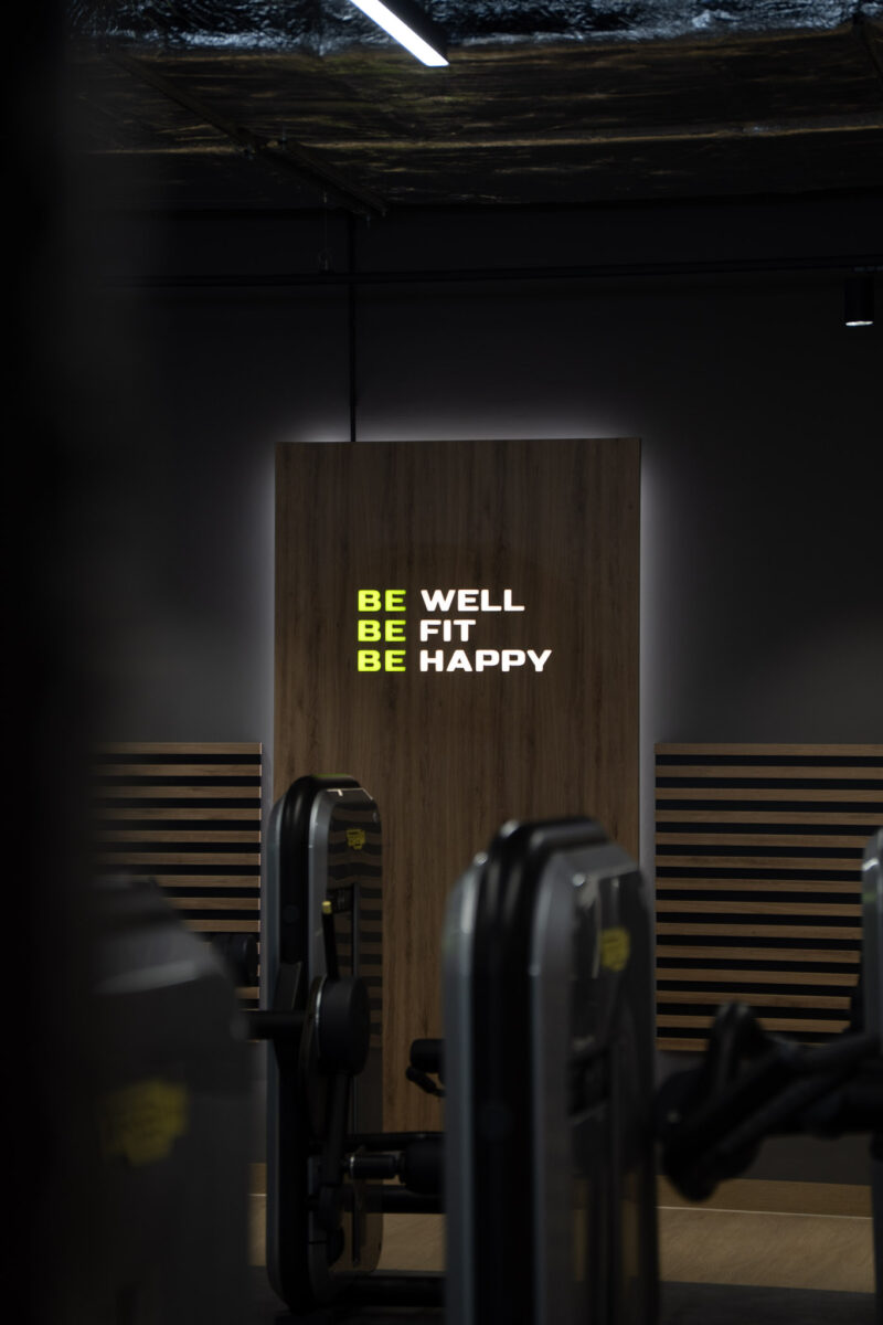 Podświetlany napis motywacyjny na drewnianym panelu: „BE WELL, BE FIT, BE HAPPY” wewnątrz klubu Well Fitness.