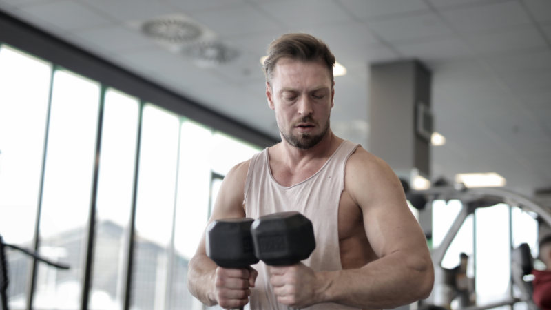 Mężczyzna w jasnej koszulce wykonujący ćwiczenia z hantlami na siłowni, co nawiązuje do wpływu testosteronu na wyniki w sporcie